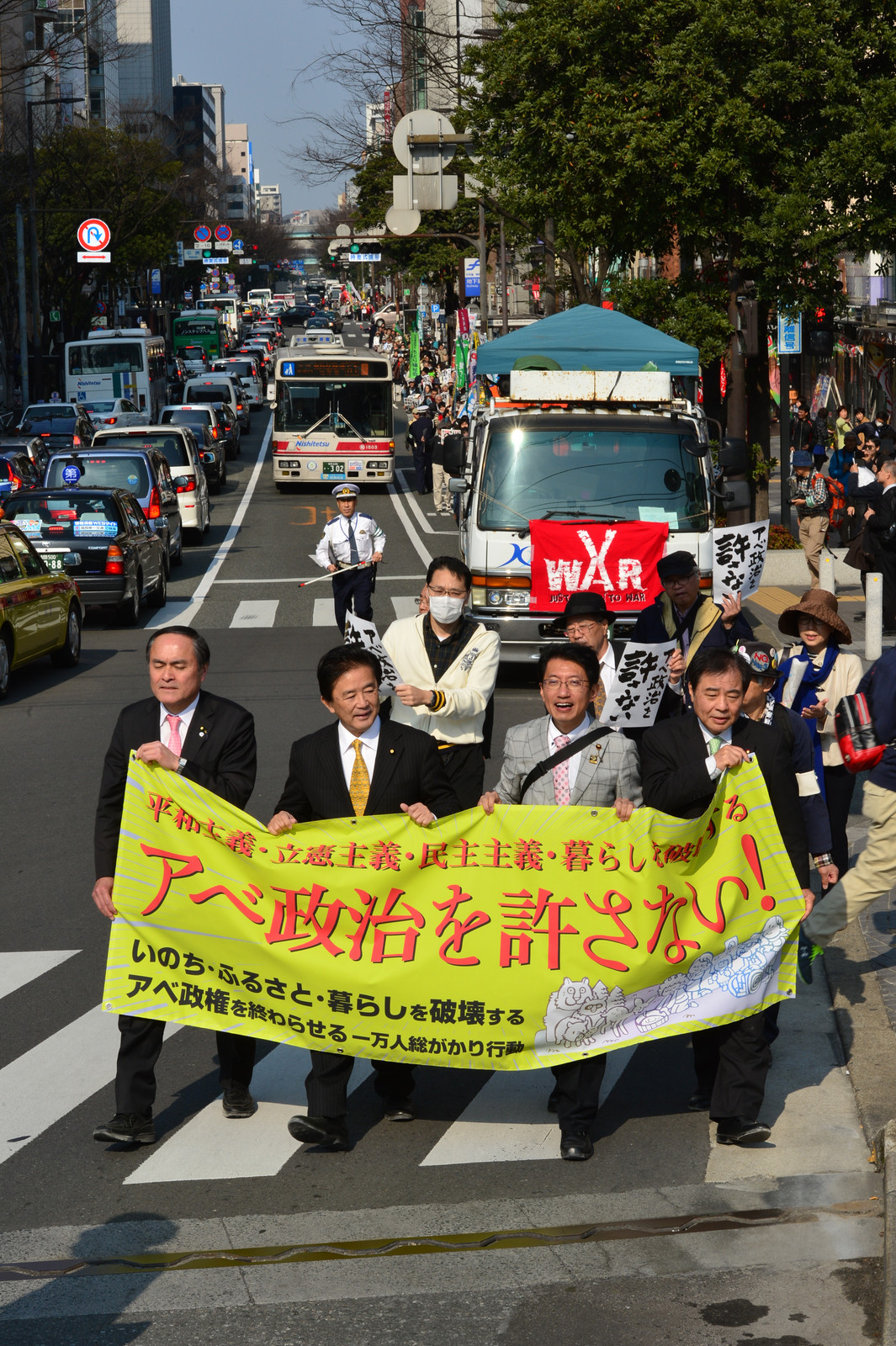 （横断幕左から）吉田、伊波、田村、荒木各氏を先頭にデモ行進する参加者ら＝19日、福岡市